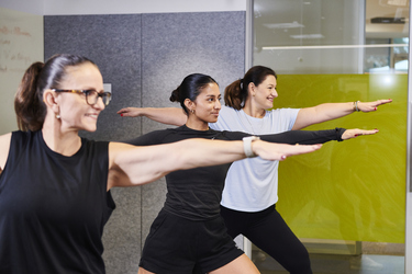 Three women stretch into warrior pose during office yoga break.