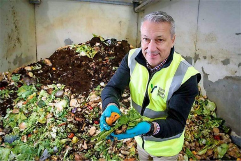 Municipal worker in safety vest holding collected organic waste, including lettuce and an orange piece, in front of a large compost pile.