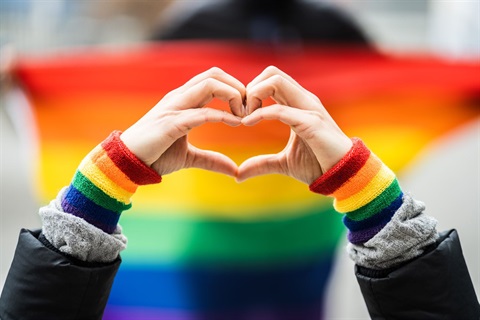 Hands formed into a heart symbol framed against the vibrant stripes of a rainbow flag.