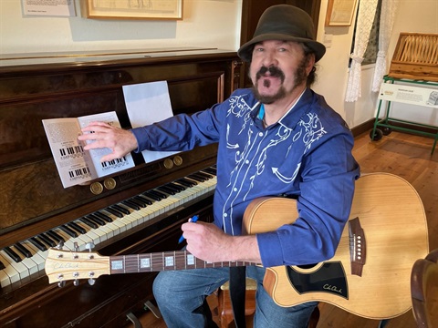 Ian Marks with songwriting booklet, sitting at a piano and holding an acoustic guitar