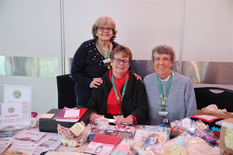 Three smiling ladies in green lanyards display handmade crafts like yarn projects and papers at an event table.