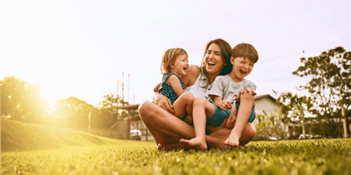 A parents sits on the grass with two young children. They are all laughing.
