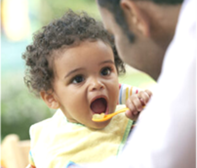 An adult watches as a baby spoons food into their mouth.