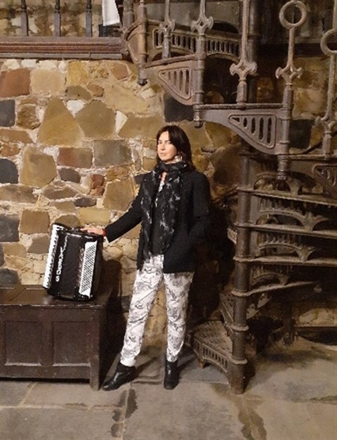A musician in a historic setting. The woman, holding an accordion, is framed by the stone walls and a grand spiral staircase. A moment of quiet contemplation.